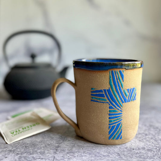 Brown ceramic mug with blue cross design on a gray surface with a blurred teapot in the background.