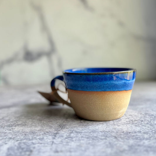A handcrafted ceramic soup mug with a blue upper rim and beige lower portion, sitting on a marble surface.