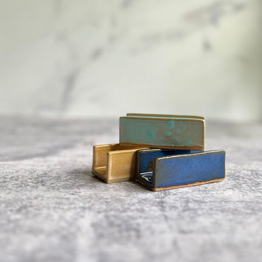 Three ceramic business card holders, one yellow, one gr, and one blue, on a marble surface.