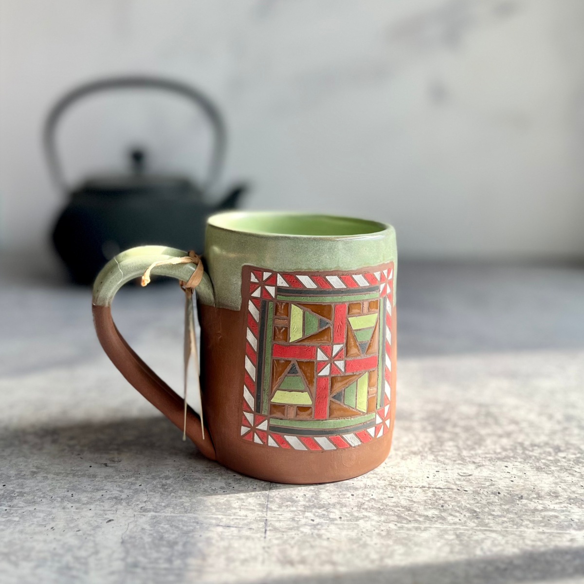 Hand-thrown ceramic mug with Christmas barn quilt pattern hand painted, green glaze on inside and upper 1/3 of outside, on chocolate brown clay. Left facing, a tea kettle in the back.