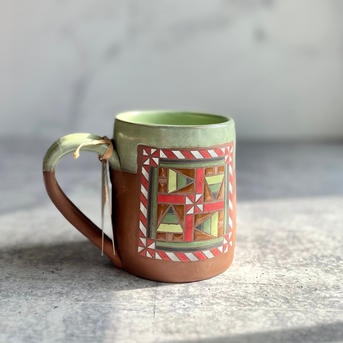 Hand-thrown ceramic mug with Christmass barn quilt pattern hand painted, green glaze on inside and upper 1/3 of outside, on chocolate brown clay. Left facing,