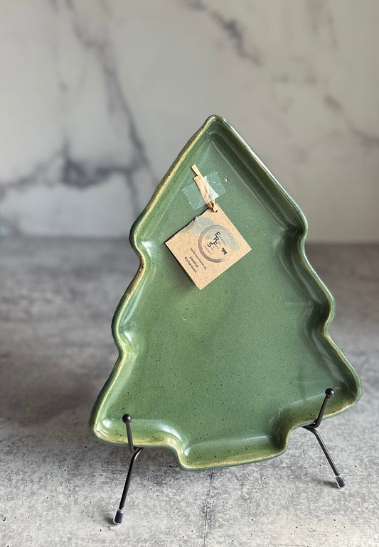 A handcrafted, green Christmas tree-shaped ceramic plate with a tag attached, displayed on a stand against a marble background.