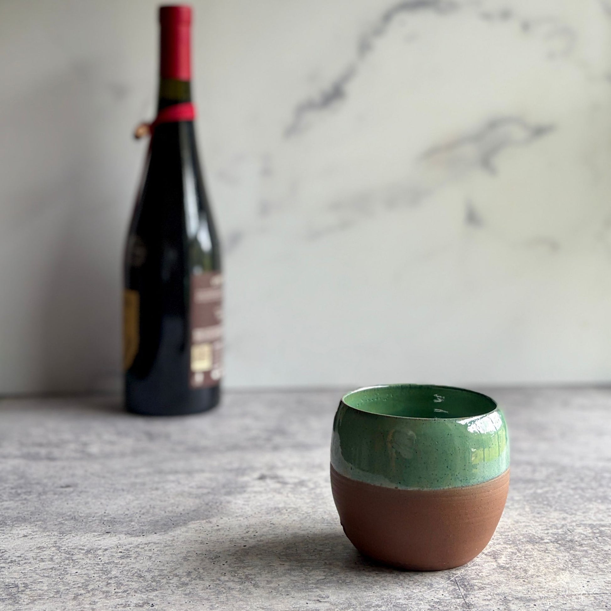 Handmade 12 oz green stoneware stemless wine cup with half-glaze design and natural clay base, wheel-thrown at End of the Road Farm Studio Alabama, with wine bottle