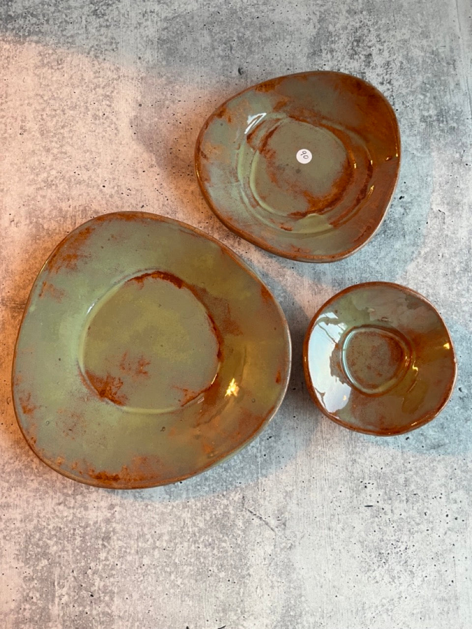 A set of three handcrafted stoneware pottery bowls in green, blue, and orange colors, with a whimsical and rustic design, displayed in a stacked arrangement on a concrete surface.
