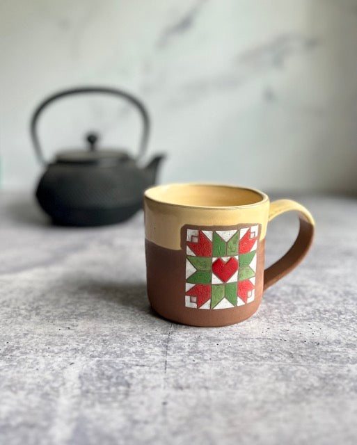 Handmade ceramic barn quilt mug with a geometric pattern of tulips and heart on a textured surface with a teapot in the background.