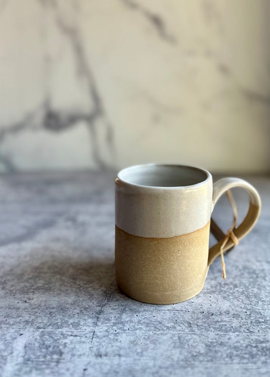 A handcrafted ceramic mug with a beige lower half and a white upper half, with a handle on the right side, placed against a marble background.