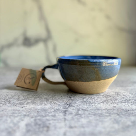 A handcrafted ceramic soup mug with a blue glaze, seated on a marble surface with a visible tag attached to it.