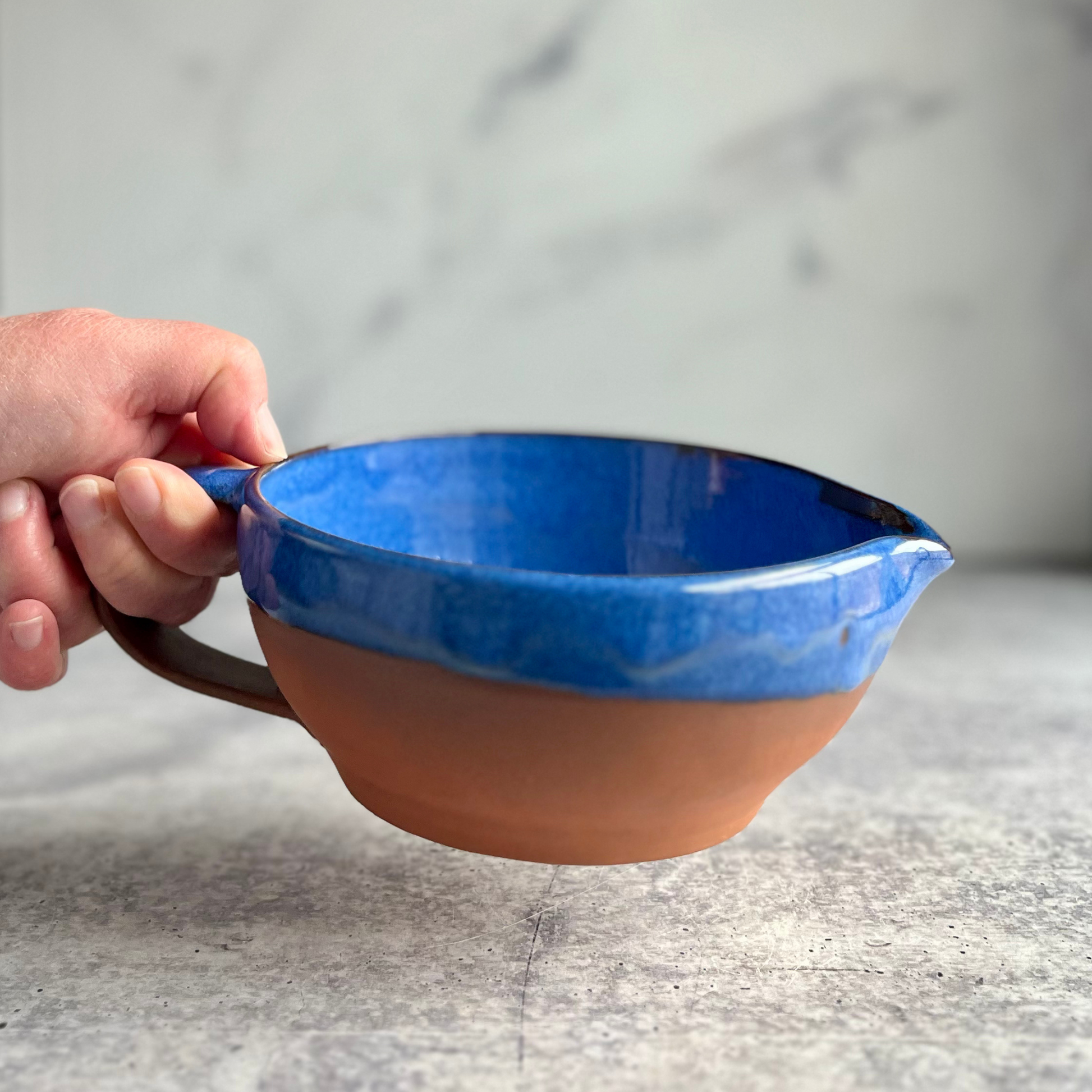 Ceramic bowl with blue interior and brown exterior held by a hand on a gray surface.