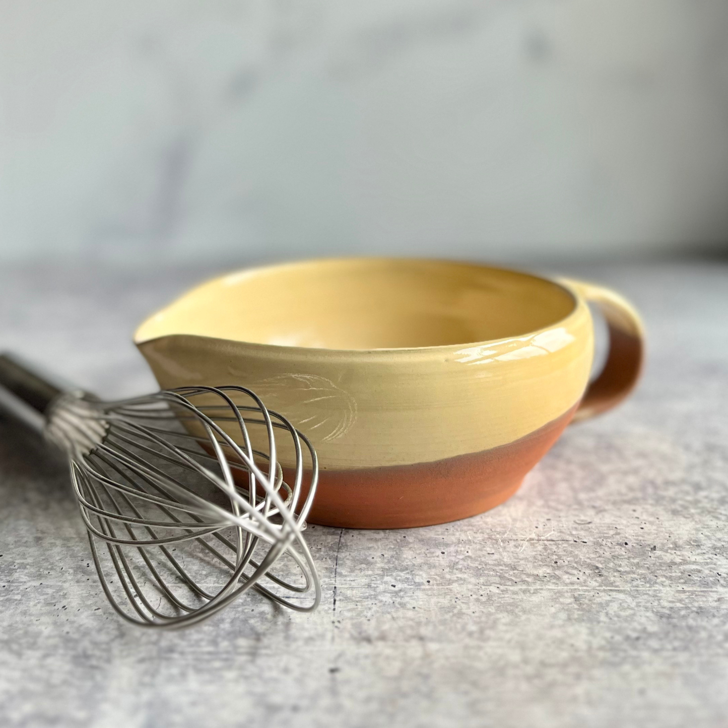 Yellow ceramic mixing bowl with a brown base and handle, accompanied by a whisk on a gray surface.