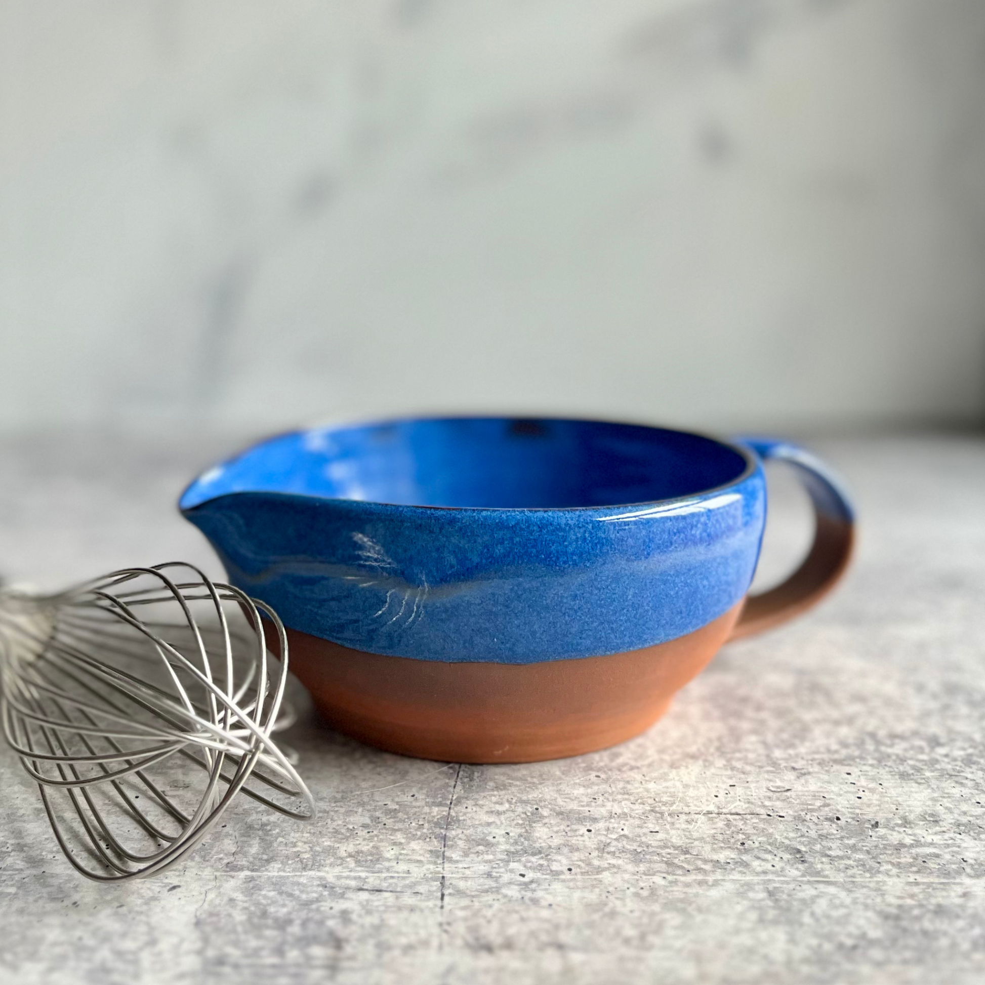Blue ceramic mixing bowl with brown base on a gray surface, next to a whisk.