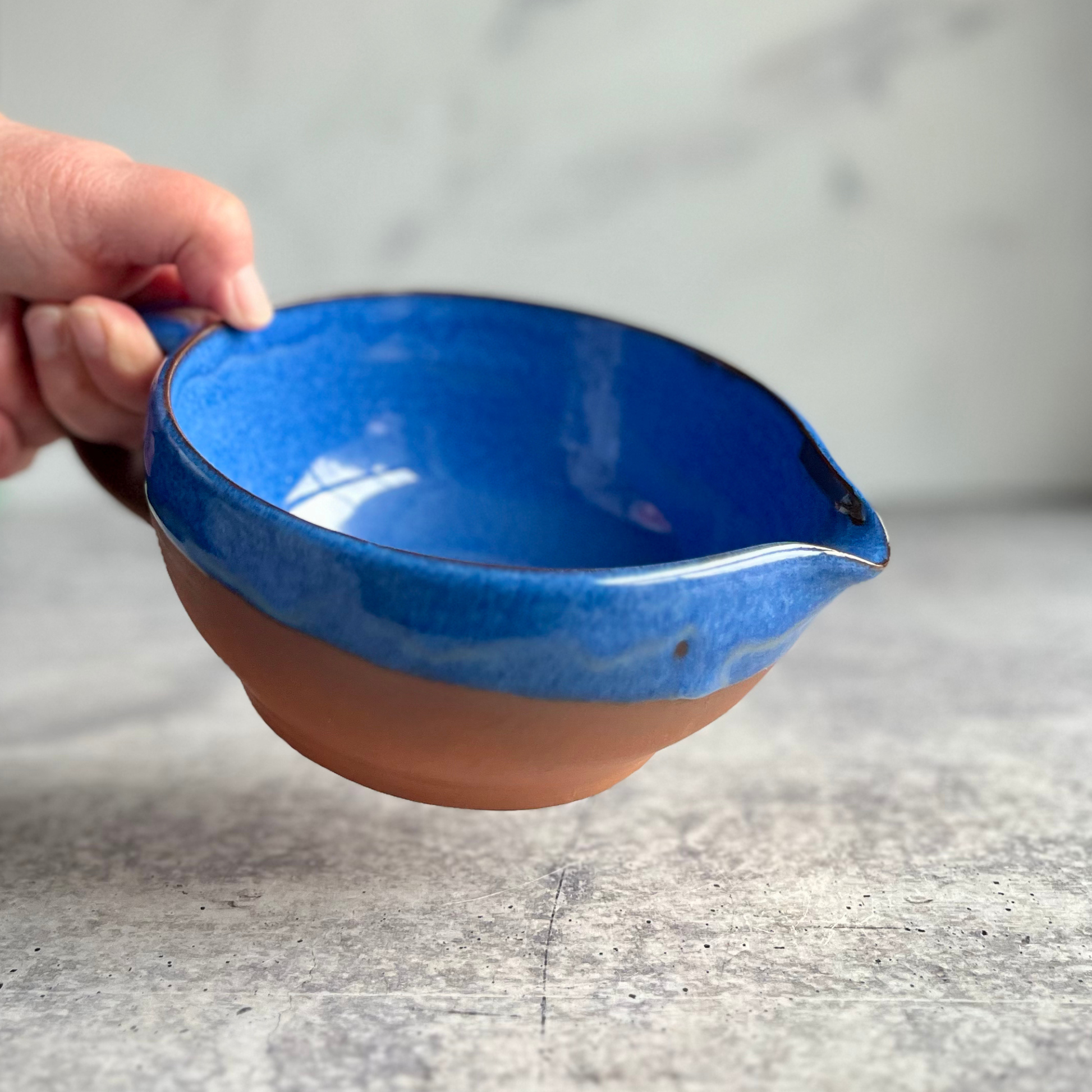 Ceramic bowl with blue exterior and brown interior held by a hand on a gray surface.