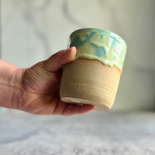 A hand holding a handmade ceramic cup with a beige base and turquoise & white glaze.