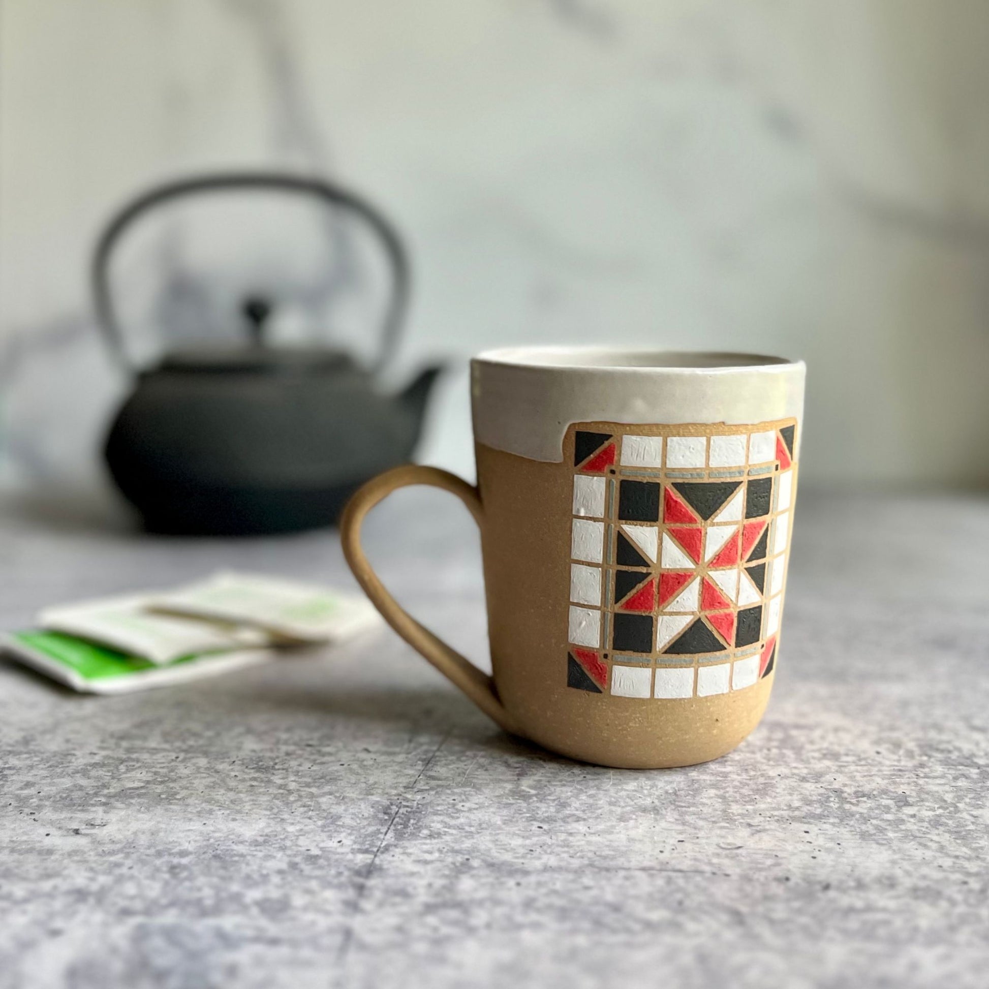 Ceramic mug with geometric pattern on a marble surface, with a teapot in the background.