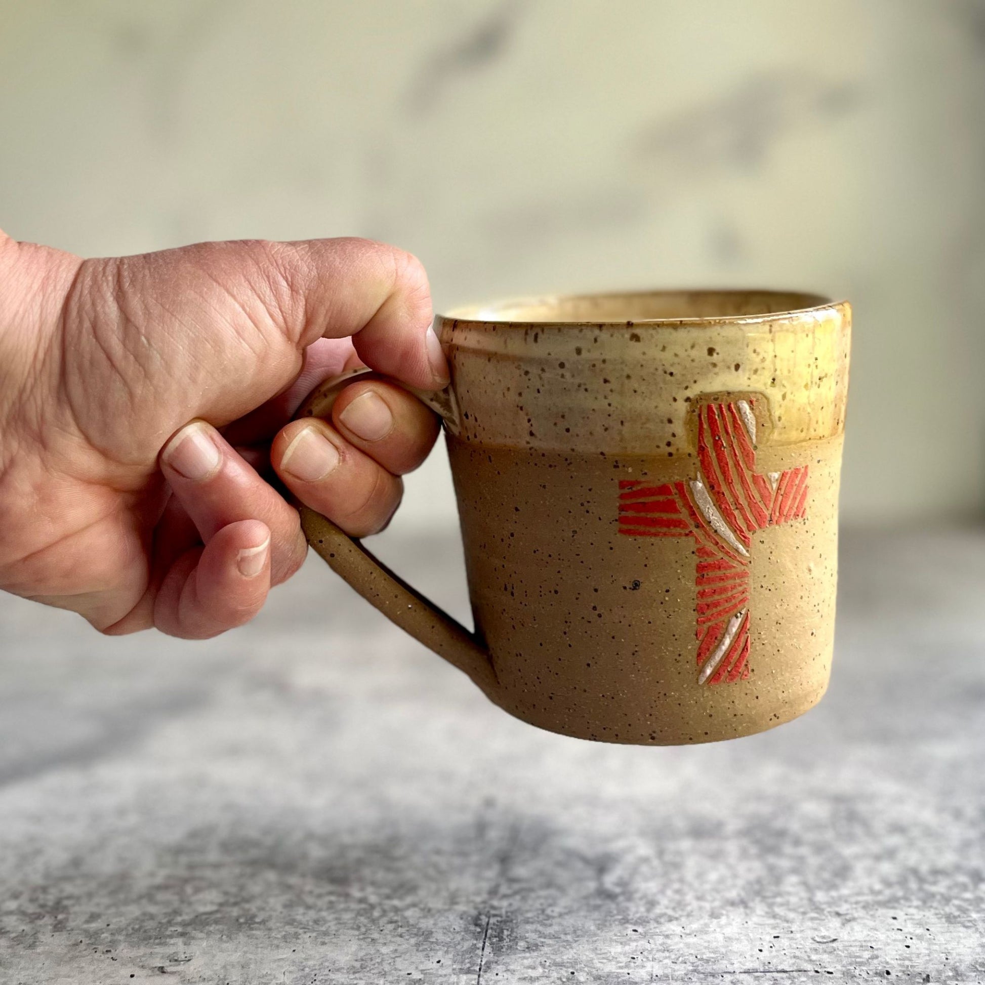 Hand holding a brown ceramic mug with a red design on a textured surface