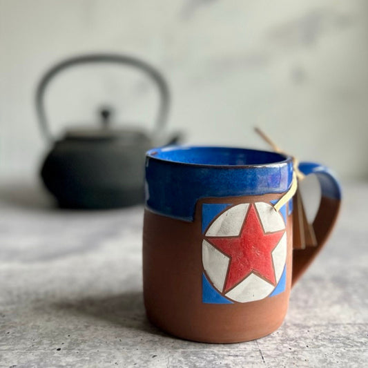 Ceramic mug with Texas star design on a textured surface, with a teapot in the background.