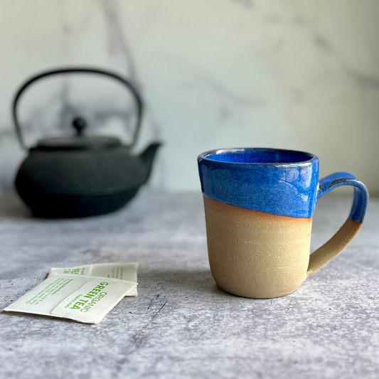 Ceramic mug with blue top and beige bottom on a marble surface with a teapot in the background.