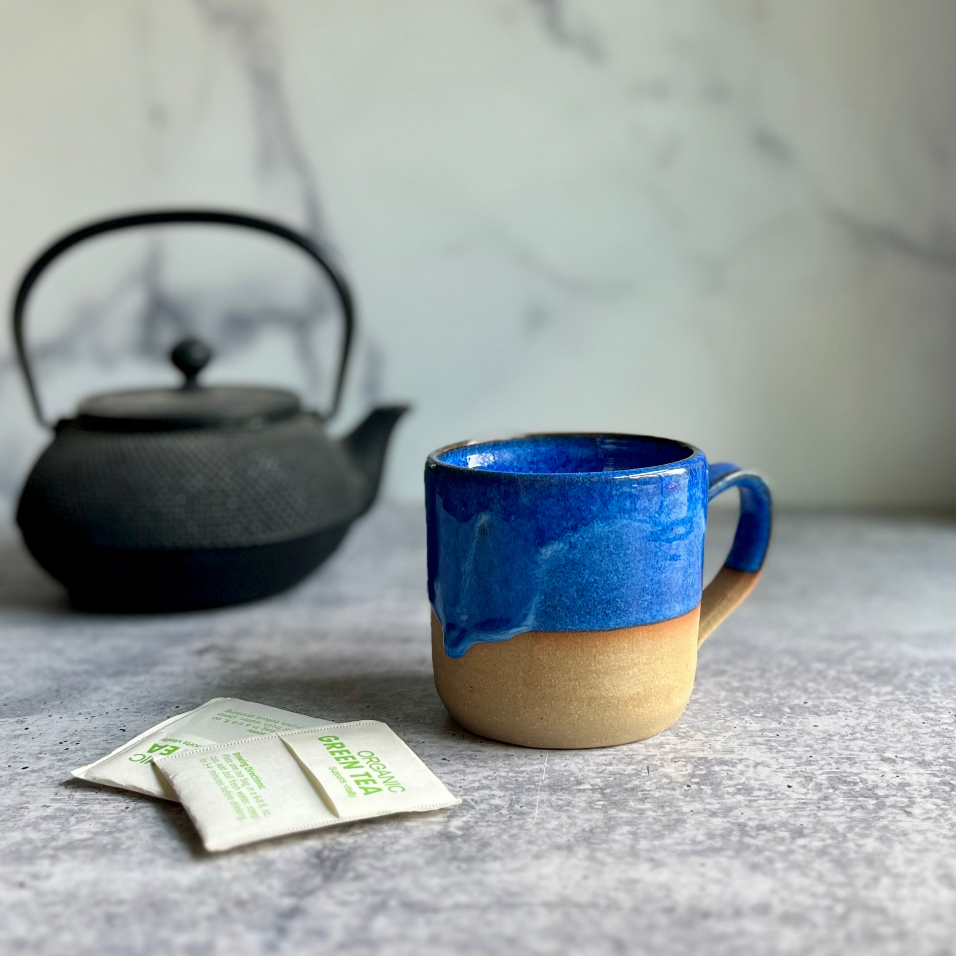 Blue ceramic mug with beige base on a marble surface with a black teapot and tea bags.