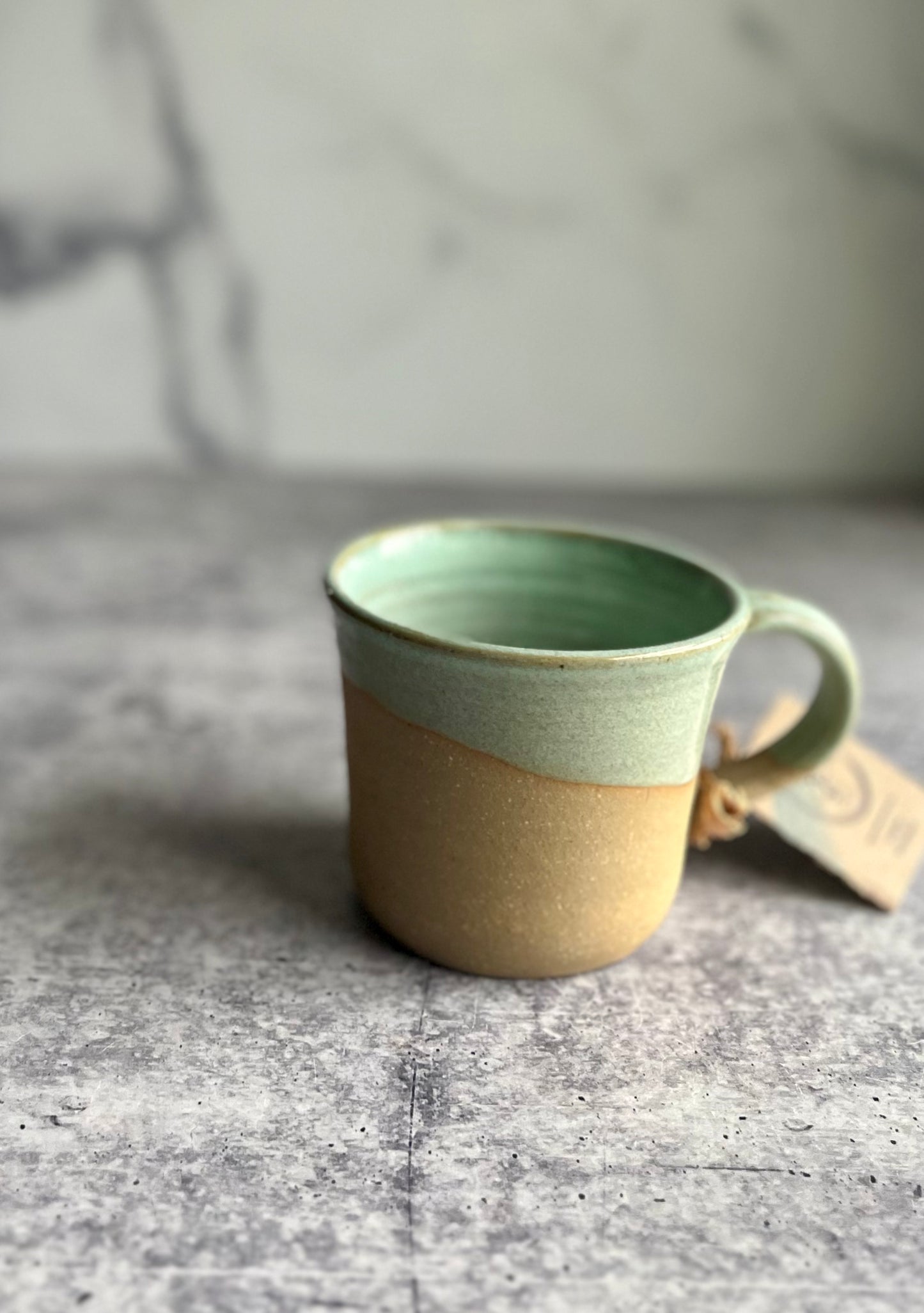 A handcrafted ceramic mug with a green upper half and beige lower half, with a tag attached, displayed against a marble background.