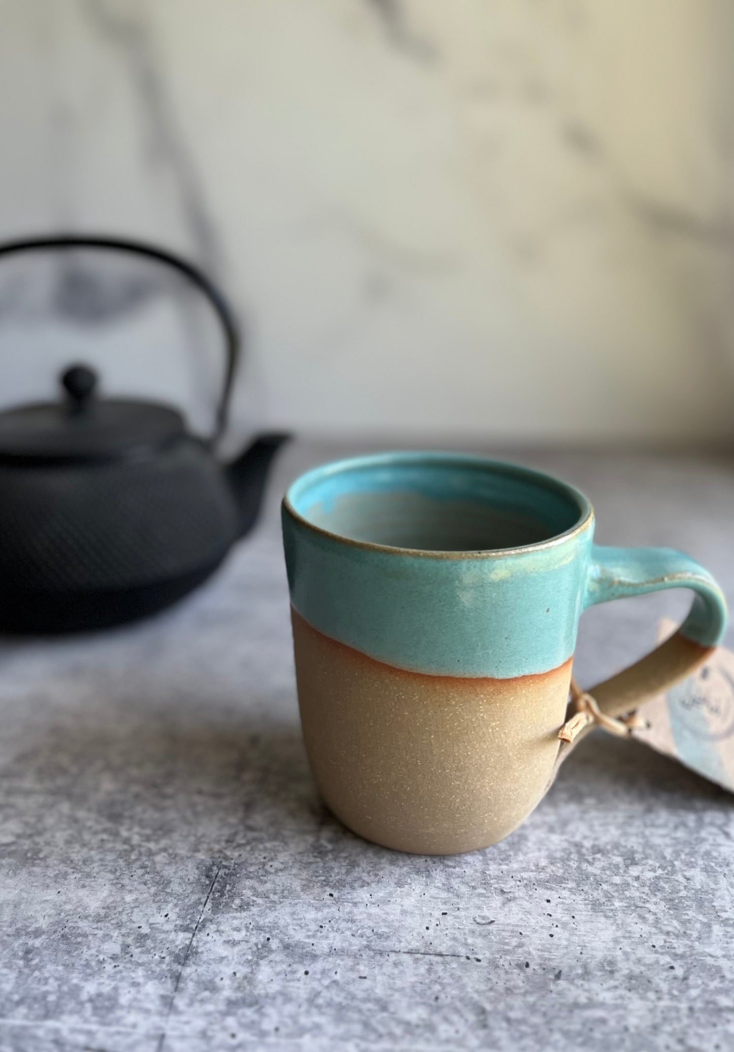 A handcrafted ceramic mug with a turquoise upper half and beige lower half, with a tag attached, displayed against a marble background.