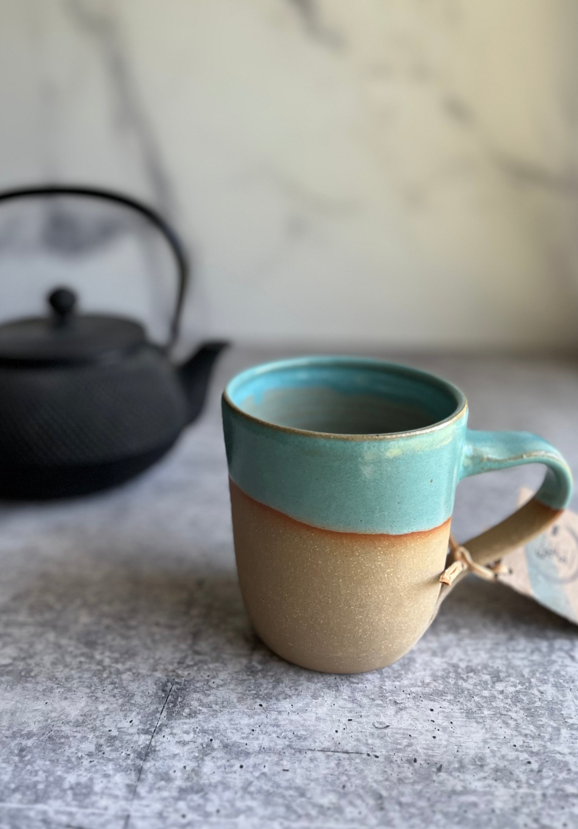 A handcrafted ceramic mug with a turquoise upper half and beige lower half, with a tag attached, displayed against a marble background.